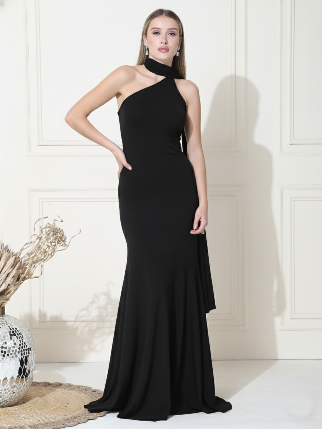 Woman wearing a black one-shoulder dress against a white wall.